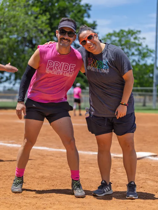 Two players on a kickball field are smiling and posing together, both wearing sporty clothing with "Pride Sports USA" shirts, athletic shorts, and sunglasses. The sunny outdoor setting features a dirt field and trees in the background.