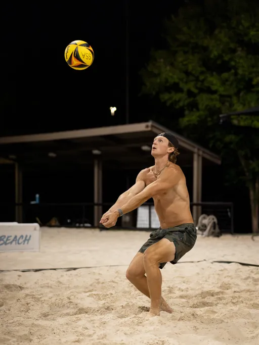On a lit sand volleyball court at night, a player in dark shorts and a backward cap bends their knees and brings forearms together to receive a yellow‑and‑black ball descending overhead; a pavilion and trees frame the background.