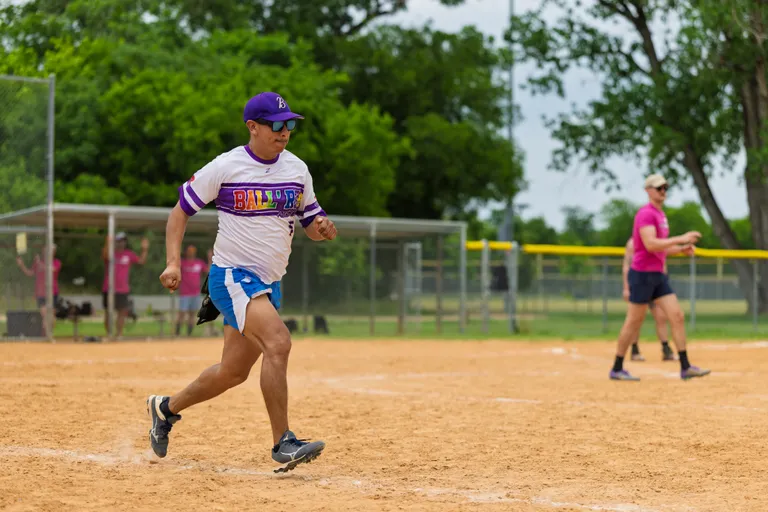 A player wearing a purple and white jersey, blue shorts, and a purple cap is running across a sandy kickball field, while another player in a pink shirt and dark shorts stands in the background ready to field.
