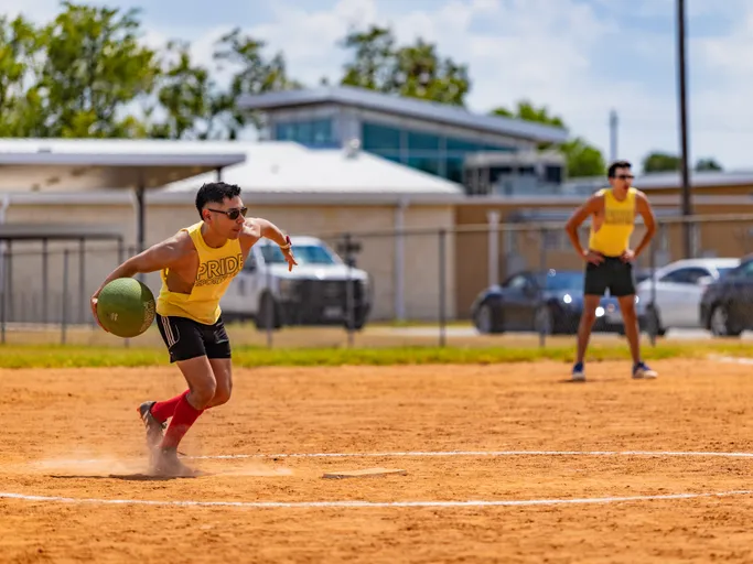 On a sunny kickball field, a Pitch Please player in a yellow “PRIDE” tank, black shorts, and red socks winds up at the pitcher’s mound with a green kickball, while a teammate in matching gear stands ready in the infield; a chain-link fence, parked cars, and low buildings sit in the background.