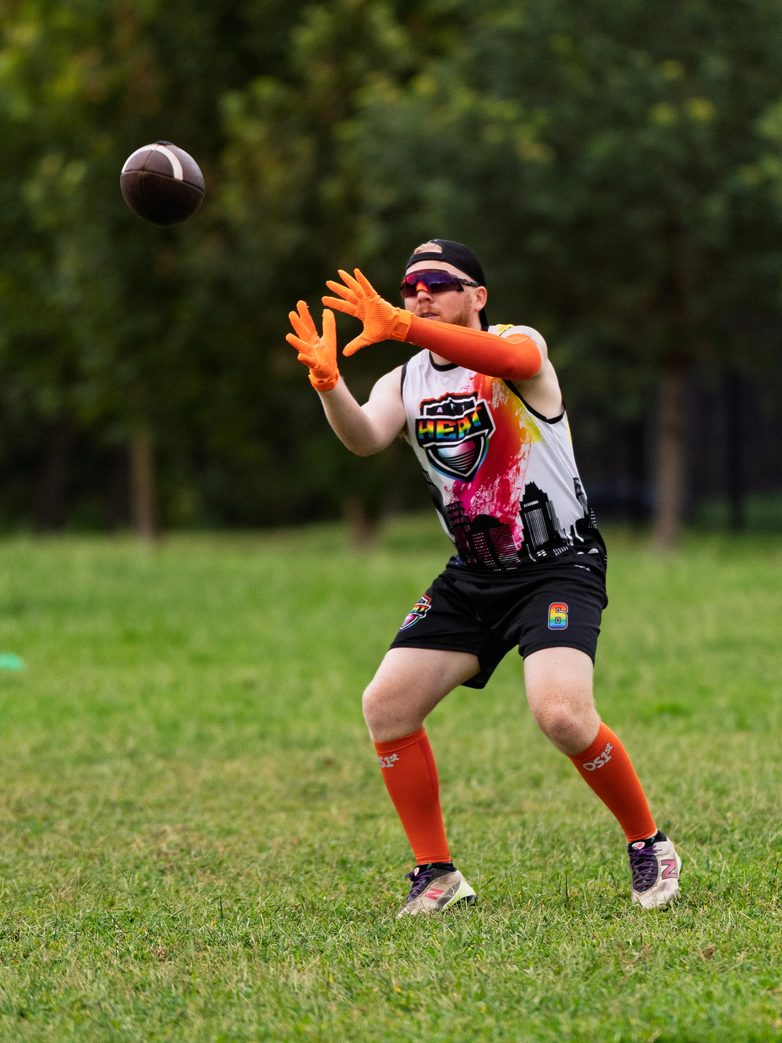 On a grassy park field, an ATX Heat (Orange) player reaches to catch a football during Gay Bowl practice, wearing a white “HEAT” team tank with cityscape graphic, black shorts, bright orange gloves and knee-high socks, a black cap, and sunglasses, with trees blurred in the background.