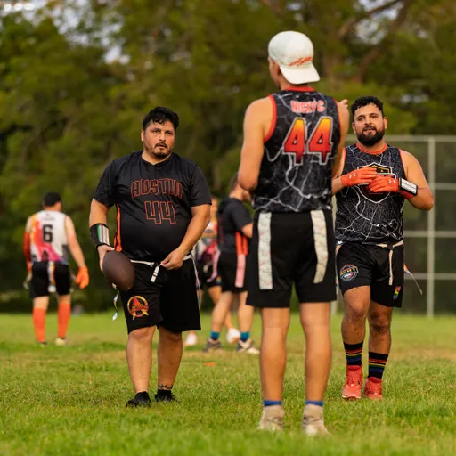On a grassy field, ATX Heat (Orange) flag football players pause mid-practice for Gay Bowl 2025, with one athlete holding a football while others in black-and-orange “Austin 44” jerseys and flag belts confer. Additional teammates move in the background, wearing gloves, cleats, and patterned uniforms.