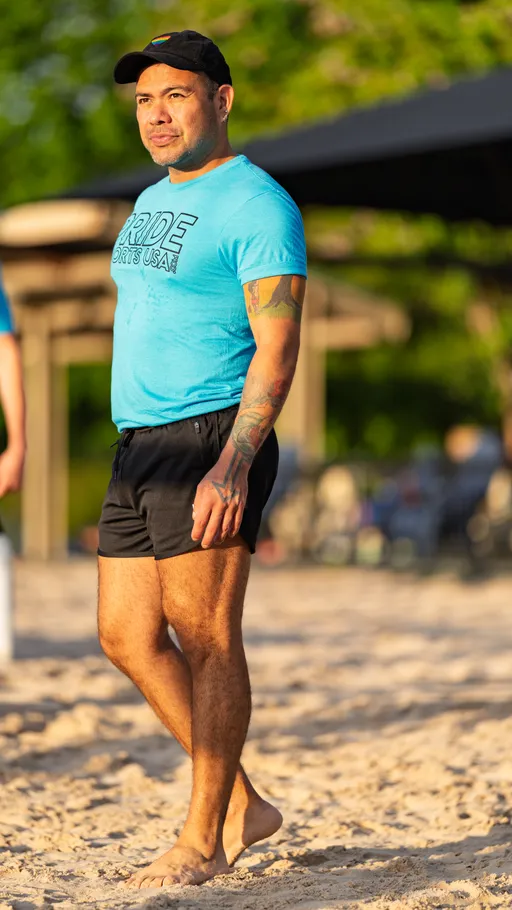 A volleyball player stands barefoot on a sand court, wearing a blue "PRIDE SPORTS USA" t-shirt, black athletic shorts, and a black cap, with trees and shaded seating visible in the background.