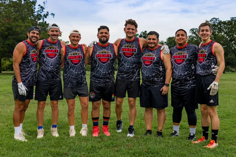 ATX Heat (Orange) players stand arm-in-arm on a grassy park field during Gay Bowl 2025 practice, wearing matching black-and-gray lightning-pattern HEAT jerseys with red trim, athletic shorts, colorful cleats, and some receiver gloves; trees and open sky form the background.