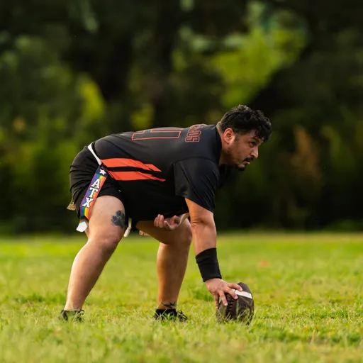 On a grassy field, an ATX Heat (Orange) player in a black-and-orange uniform crouches at center, gripping the football to snap during practice, wearing cleats and a forearm sleeve with “ATX” text visible on the shorts and trees blurred in the background.