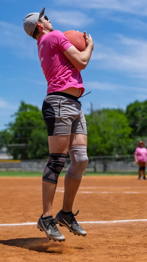 A player wearing a pink shirt, gray shorts, knee braces, and a cap is jumping in the air while holding a red kickball close to their chest on a kickball field, with another player visible in the background.