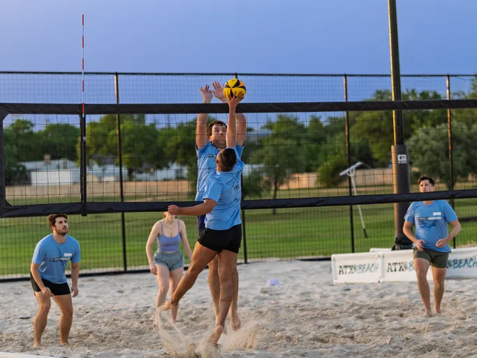 Two volleyball players wearing blue shirts are jumping at the net, reaching for the ball, while teammates stand ready behind them on a sandy outdoor sand volleyball court surrounded by a fence and green field.