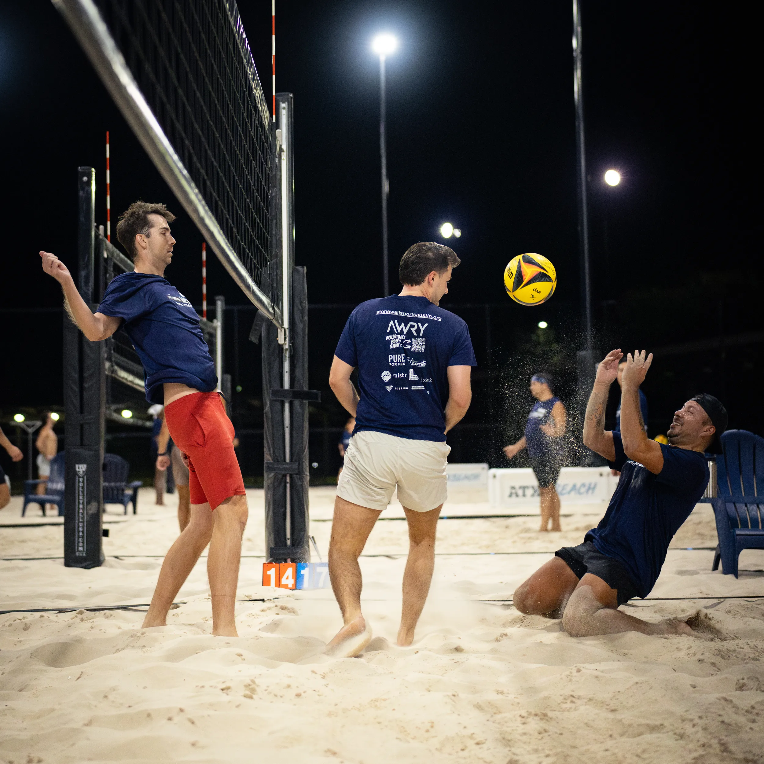 Night sand volleyball action: three players in navy shirts react as a yellow-and-black ball drops near the net—one jumping by the pole, one bracing in tan shorts, and one falling backward into the sand with hands up to make a play. The scene is lit for night play with multiple courts and blue chairs visible in the background.