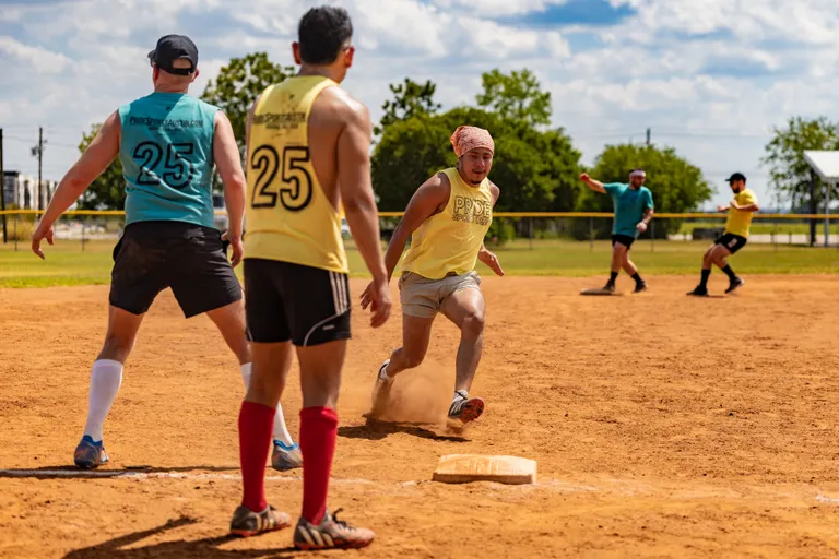 On a sunny kickball field, a player in a yellow jersey sprints through a base as dust kicks up, while teal- and yellow-clad defenders move in; numbers, knee-high socks, and headbands stand out against a grassy outfield and chain-link fence.