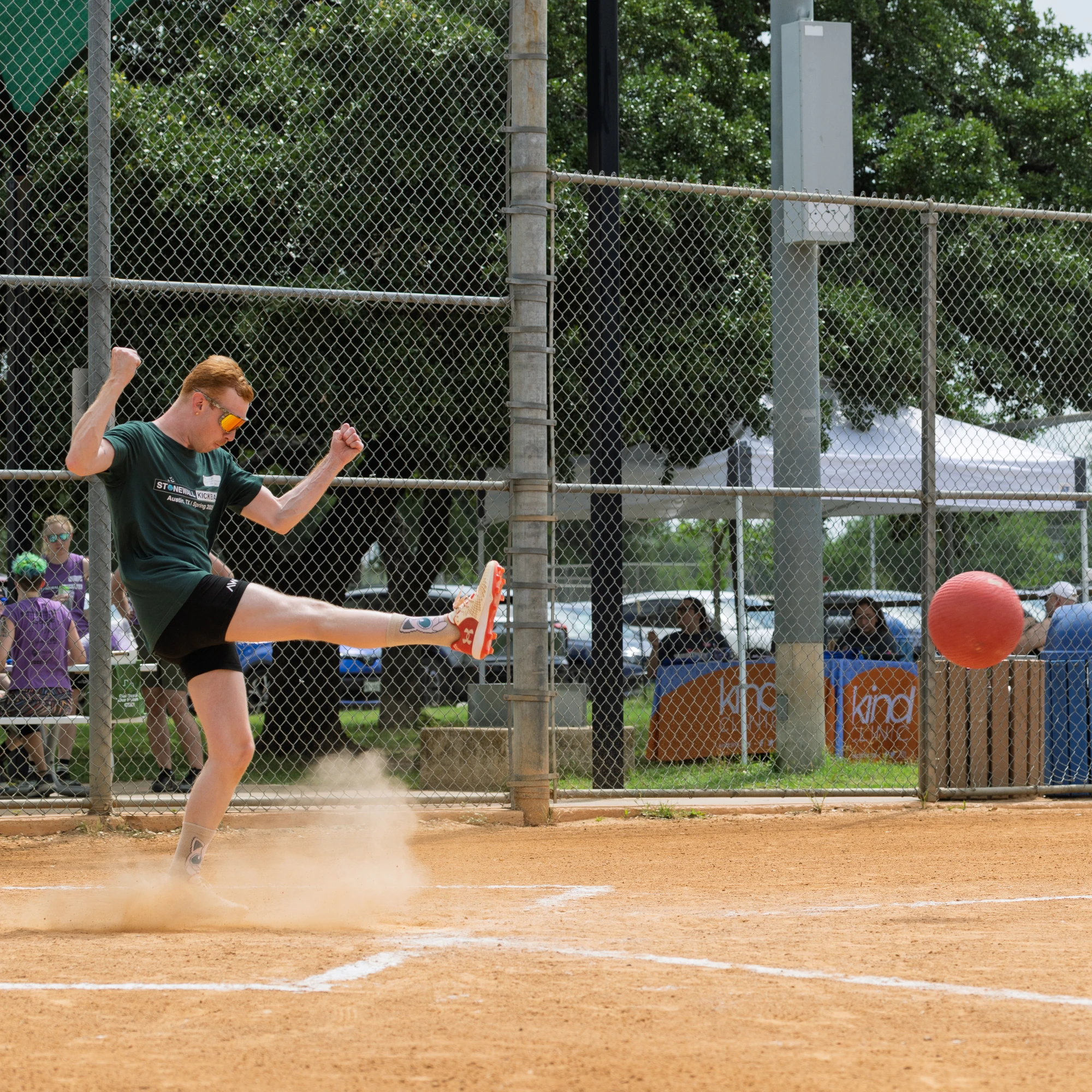 A player wearing a dark athletic shirt, shorts, and sunglasses is kicking a red kickball on a dirt field, with a cloud of dust rising from their foot and a chain-link fence and spectators in the background.
