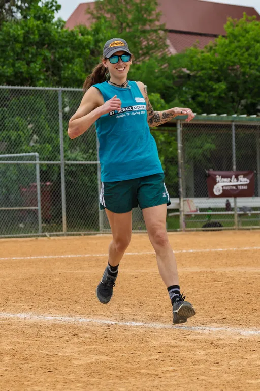 A player wearing a teal Stonewall Kickball tank top, dark athletic shorts, sunglasses, and a cap is running on a kickball field, appearing to celebrate or cheer with thumbs up. A chain-link fence and greenery are visible in the background.