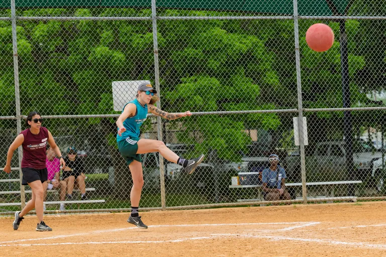 A player in a teal athletic jersey and shorts is kicking a red kickball on a dirt field, while another player in a maroon jersey runs nearby; a chain-link fence and green trees are visible in the background, with a few people seated on benches.