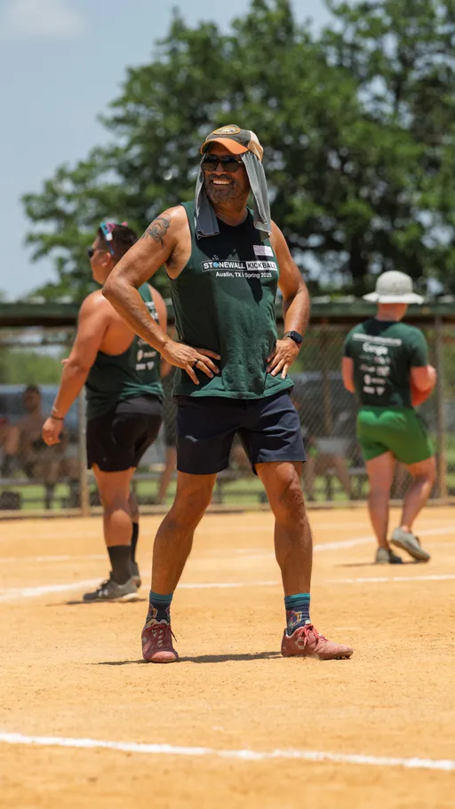 A kickball player stands on the field with hands on hips, wearing a sleeveless team shirt, shorts, sneakers, sunglasses, and a cap with a cloth draped for sun protection; other players and a fence are visible in the background.
