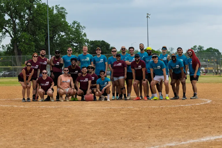 A group of kickball players wearing matching maroon and blue team shirts pose together on a kickball field, with one player holding a red kickball in front of them. The setting is outdoors on a dirt field with grass and trees visible in the background.