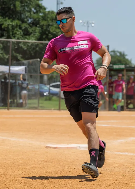 A player wearing a bright pink "Stonewall Kickball" shirt, black shorts, black knee-high socks with a pink ribbon, and blue sunglasses is running on a sandy kickball field, with other similarly dressed participants and a fence visible in the background.