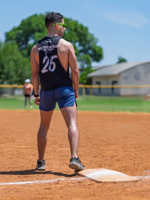 A kickball player wearing a sleeveless jersey with the number 25, blue shorts, and sunglasses stands near a base on a sunny outdoor kickball field, appearing ready for action as another player is visible in the background.