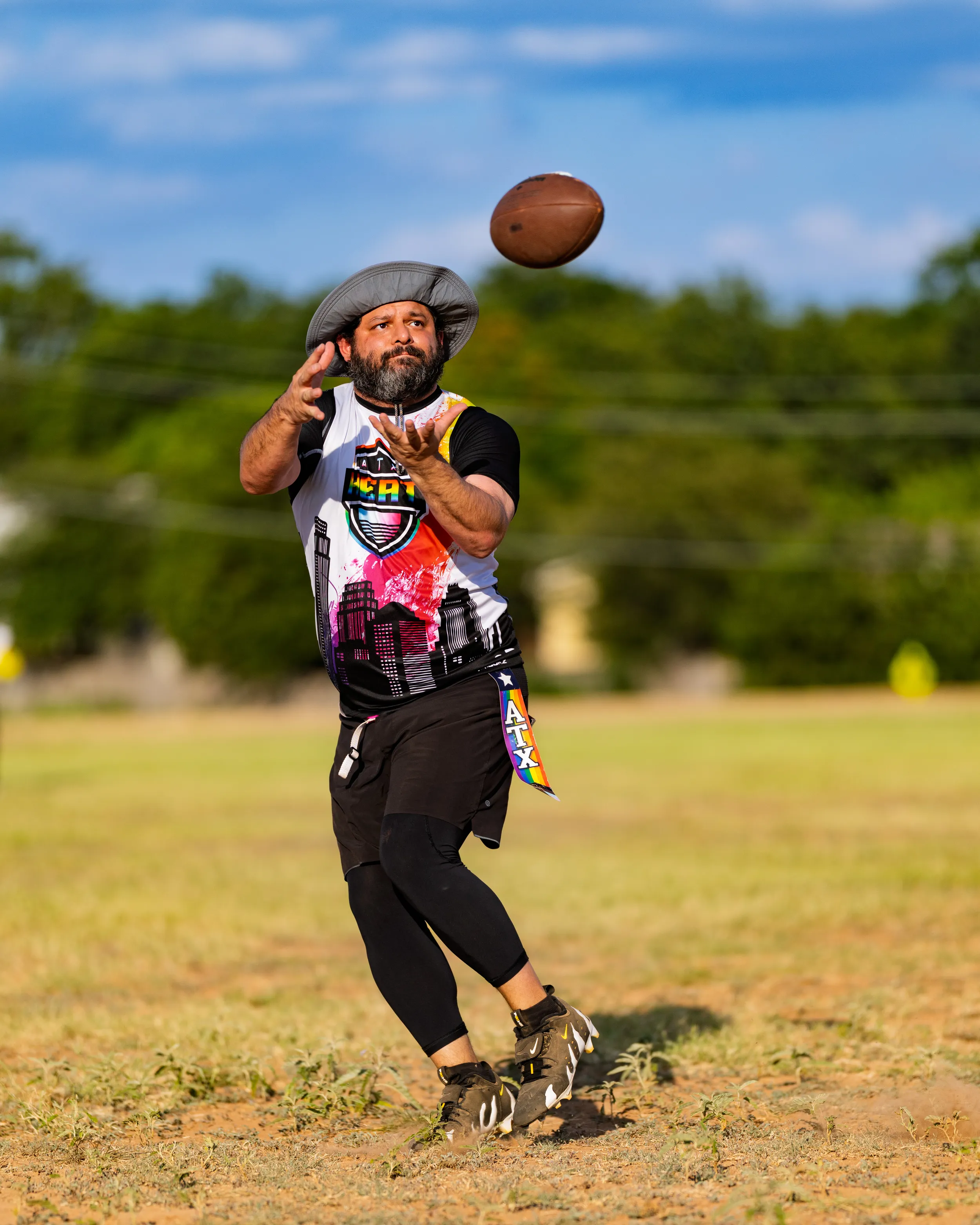 During ATX Heat (Black) practice for Gay Bowl 2025, a flag football player in a graphic ATX Heat jersey, black leggings, cleats, and a cowboy hat runs toward an incoming football with hands ready to catch, a rainbow “ATX” flag belt trailing on a sunny grassy field with trees in the background.
