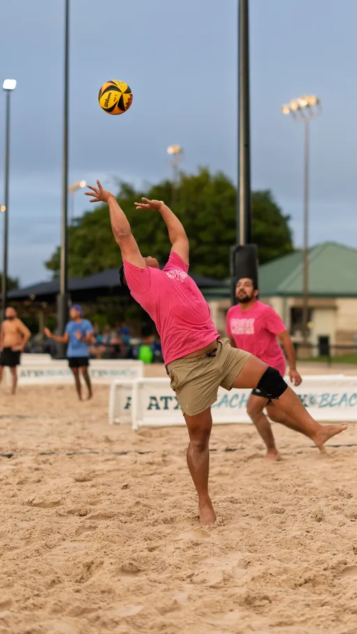 A volleyball player wearing a bright pink shirt and tan shorts jumps to hit a ball over the net on a sand court, with other athletes and volleyball nets visible in the background under evening lights.