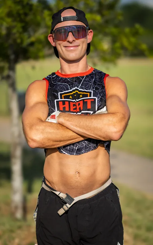 An ATX Heat (Black) flag football player stands on a grassy practice field with arms crossed, wearing a black-and-red cropped sleeveless jersey with the ATX Heat logo, black shorts with a flag belt, large sport sunglasses, and a backward cap; sunlit trees blur in the background during practice for Gay Bowl 2025.