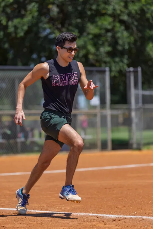 A player wearing sunglasses, a sleeveless "Pride Sports USA" shirt, shorts, and athletic shoes is running on a kickball field, with a chain-link fence and trees in the background.