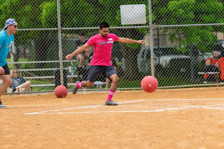 A player in a pink shirt, dark shorts, and pink striped socks is kicking a red kickball on a dirt field while another player in a blue shirt watches nearby; the scene takes place outdoors with a chain-link fence and spectators in the background.