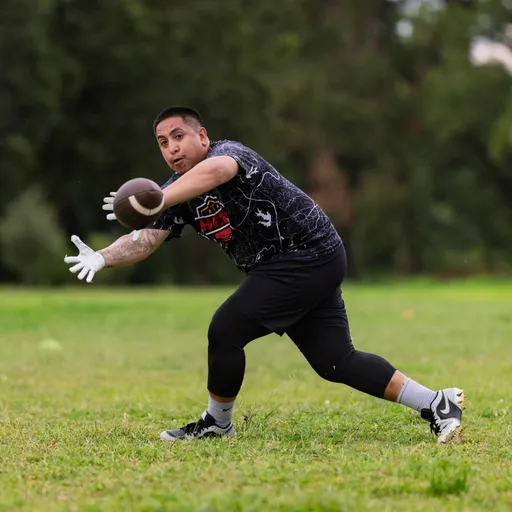 On a grassy field, an ATX Heat flag football player lunges forward with outstretched, gloved hands to snag a low pass during practice, wearing a black patterned team jersey with logo, leggings, socks, and cleats; trees blur in the background.