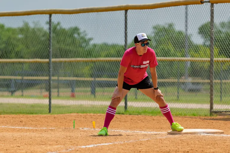 A kickball player in a pink team shirt, knee-high striped socks, neon green shoes, and a cap is in a ready stance on a dirt field near a base, preparing to make a play. The player is wearing sunglasses and the field is enclosed by a chain-link fence with grassy areas in the background.