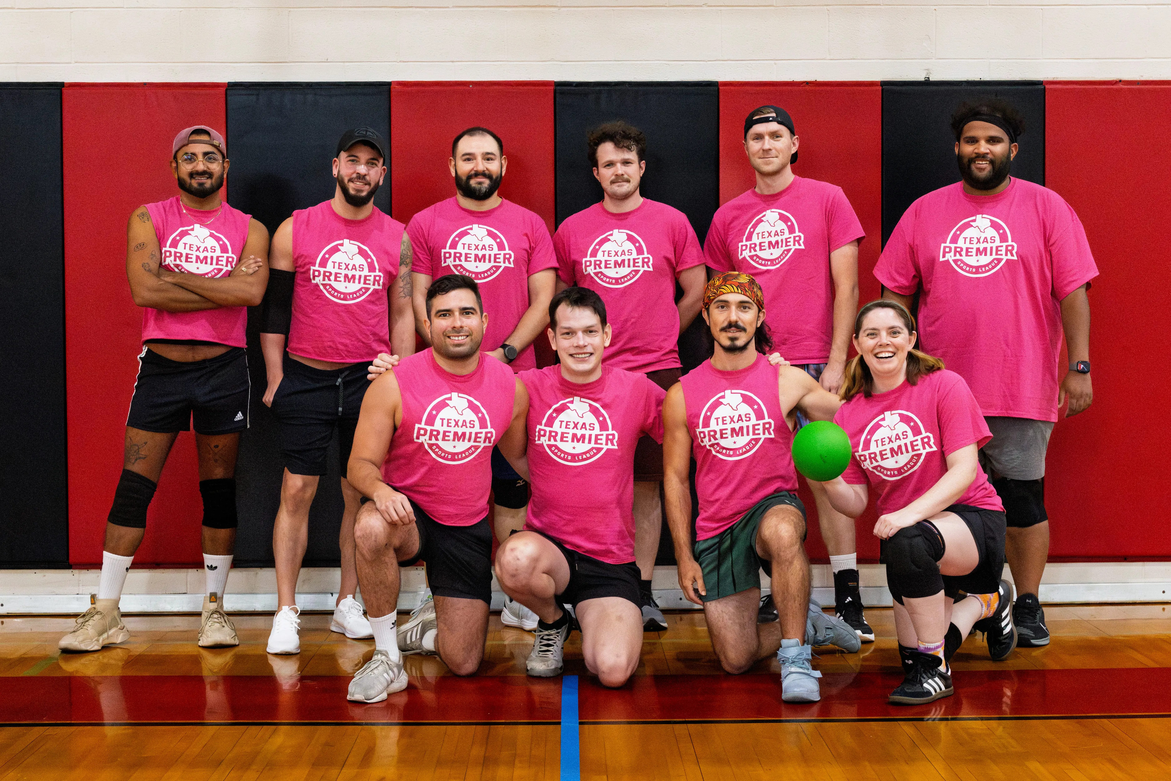 A group of dodgeball players poses for a team photo in an indoor gym, wearing matching pink “Texas Premier” shirts with athletic shorts and sneakers; some have knee pads, hats, or headbands. Several kneel in front—one holding a bright green dodgeball—against a red-and-black padded wall.