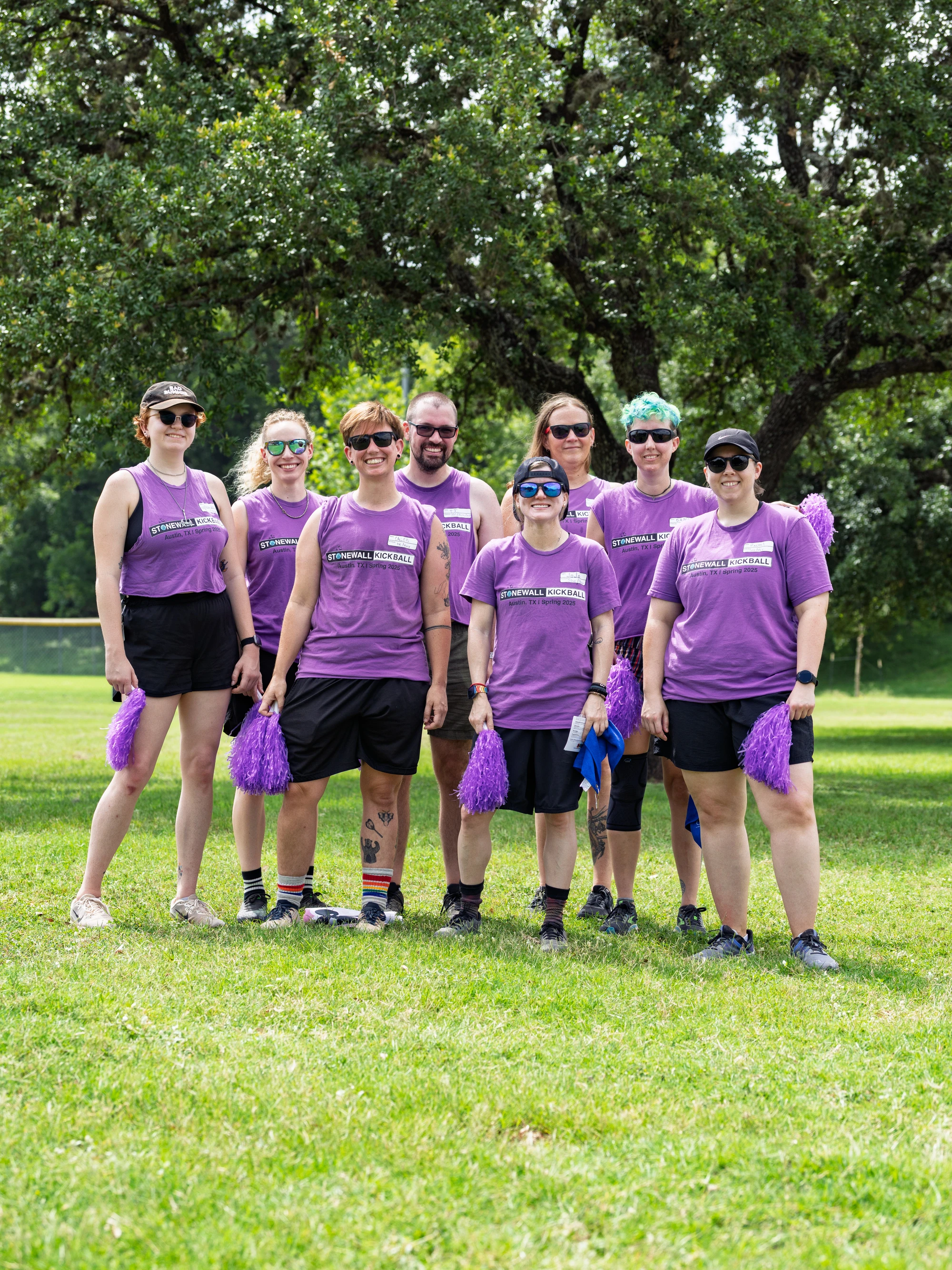 A group of eight kickball players wearing matching purple Stonewall Kickball shirts, shorts, and athletic shoes are standing together on a grassy field, holding purple pom-poms, with trees visible in the background.