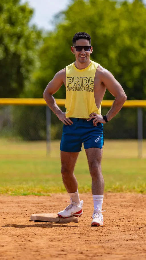 A player stands on a kickball base with hands on hips, wearing a yellow “PRIDE SPORTS USA” sleeveless top, blue shorts, white socks, red-trimmed cleats, sunglasses, and a smartwatch; the sunlit field has a dirt infield, chain-link fence, and trees in the background.