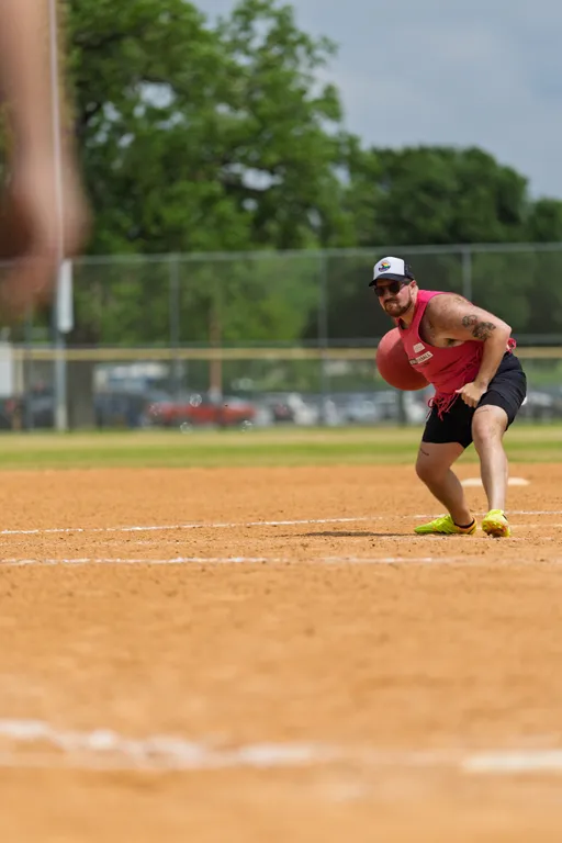 A kickball player wearing a bright tank top, shorts, a cap, and athletic shoes is preparing to throw a red kickball on a dirt field, with another player visible in the foreground and a fence with trees in the background.