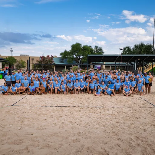 A large group of volleyball players in matching blue shirts pose together on a sandy sand volleyball court, with outdoor pavilions and trees visible in the background.