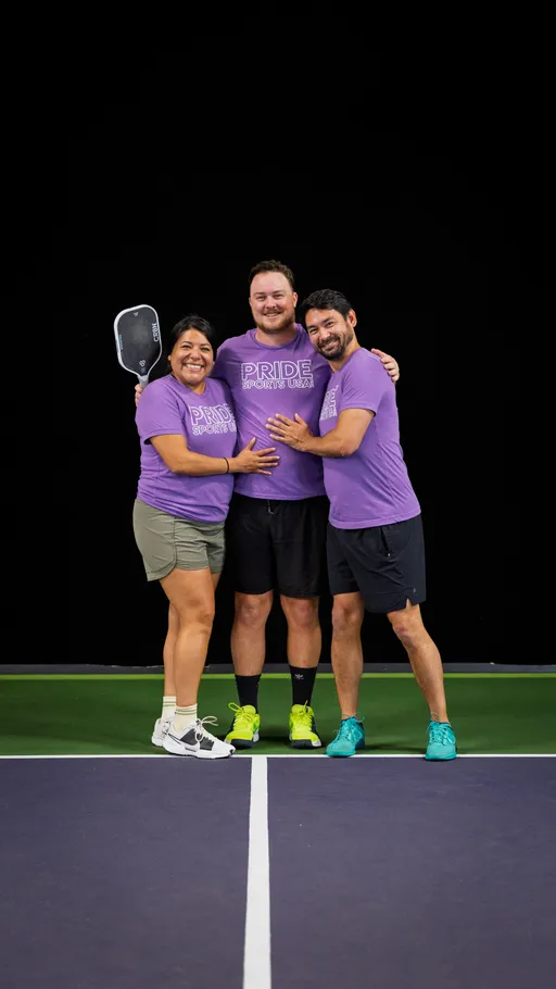 Three pickleball players stand together smiling on an indoor court, each wearing matching purple "Pride Sports USA" shirts, with one person holding a pickleball paddle.