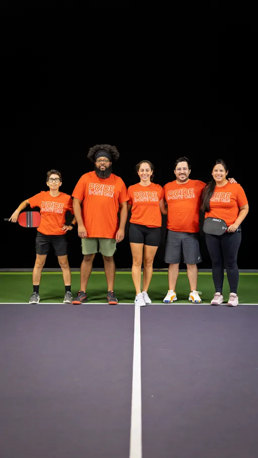 Five pickleball players stand together on a court, each wearing matching orange "Pride Pickleball 2025" shirts; two of the players hold pickleball paddles, and they are grouped in a line with arms around each other, smiling in a well-lit indoor setting.