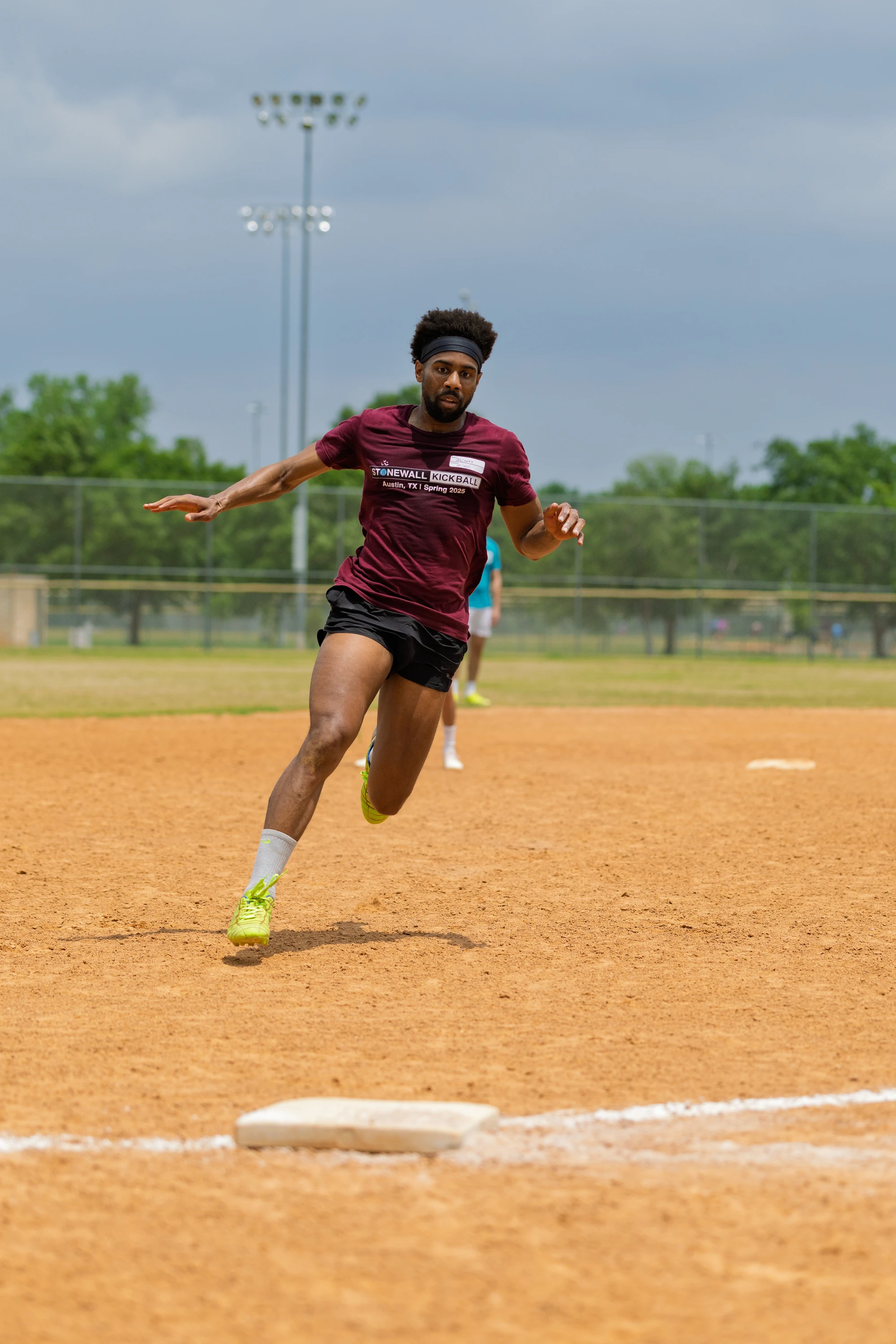 A player wearing a maroon "Stonewall Kickball" shirt, black shorts, and yellow cleats is running at full speed toward a base on a dirt kickball field, with green trees and field lights visible in the background.