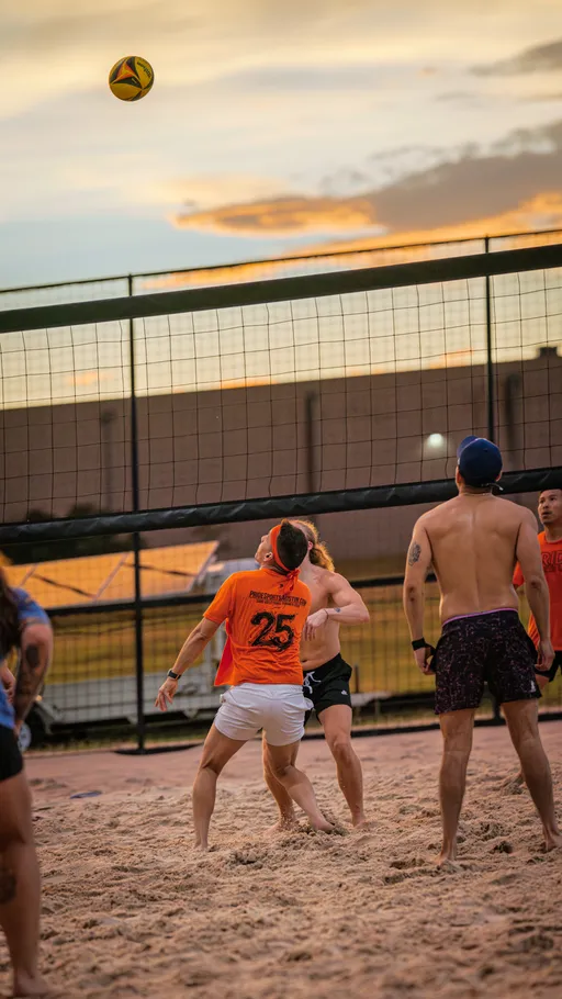 Several volleyball players stand on a sandy court at sunset, watching a volleyball in midair above the net; players wear athletic clothing, including shorts and t-shirts, with some wearing hats or visors.
