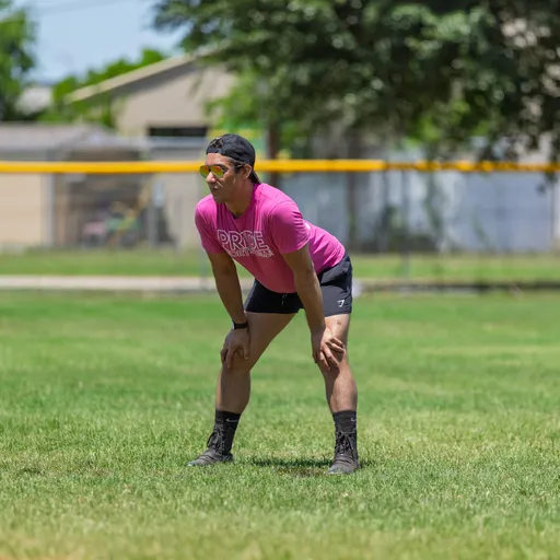 A player wearing a bright pink "Pride Kickball" shirt, black shorts, patterned socks, and sunglasses is positioned on a grassy kickball field, leaning forward with hands on knees and appearing focused on the game.