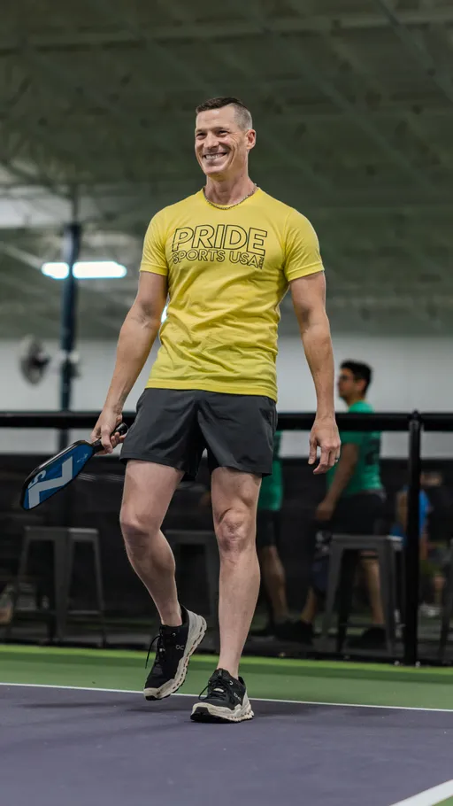 A player wearing a yellow "Pride Sports USA" shirt, black athletic shorts, and sneakers is smiling while holding a pickleball paddle on an indoor pickleball court. Other competitors and seating are visible in the background.