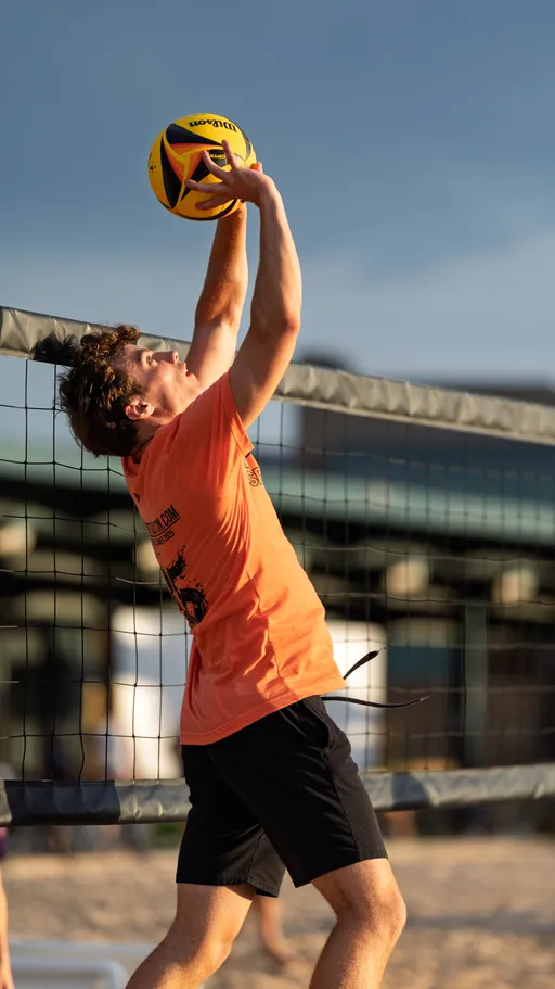 A volleyball player in an orange shirt and black shorts jumps to block or set a volleyball above their head at the net on a sand court, with an outdoor background visible behind them.
