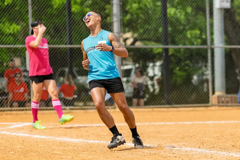 A player wearing a bright blue sleeveless shirt, black shorts, sunglasses, and sneakers celebrates on a sandy kickball field, while another player in a pink shirt, black shorts, and striped knee socks stands in the background near home plate.