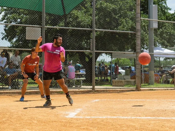 A kickball player in a bright pink shirt and black shorts is preparing to throw a kickball, while another player in an orange shirt and athletic shoes stands ready nearby on a dirt kickball field with a chain-link fence and spectators in the background.