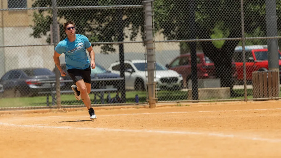 A player wearing a bright blue Stonewall Kickball t-shirt, black shorts, and sunglasses is sprinting across a sandy kickball field, with a chain-link fence and parked cars visible in the background.
