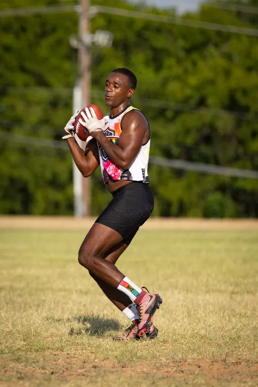 An ATX Heat (Black) player practices for Gay Bowl 2025, gripping a football with white gloves and settling into a passing stance on a sunlit grass field. The athlete wears a colorful ATX tank, black shorts, patterned socks, and cleats, with trees and utility lines in the background.