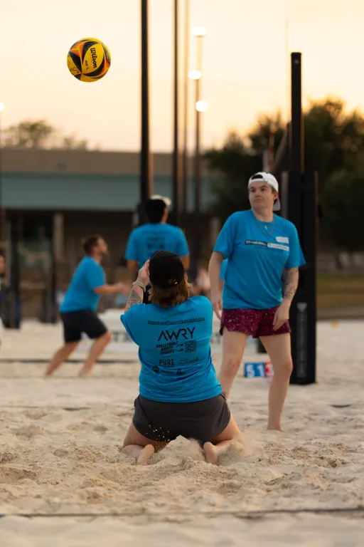During a Stonewall Sand Volleyball game at dusk, a player in a blue team shirt and cap kneels in the sand to receive a yellow-and-black volleyball dropping overhead, while a nearby teammate in patterned shorts tracks the play near the net. Other players, multiple nets, and a small scoreboard are visible in the background, emphasizing active league play.