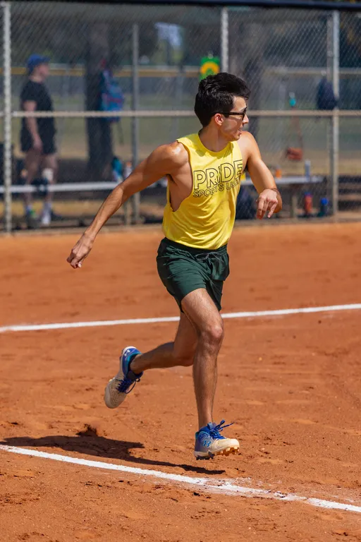 A kickball player in a yellow “PRIDE SPORTS” tank, dark green shorts, blue sneakers, and sunglasses runs along a white-chalk baseline on a red-dirt kickball field, looking toward the infield. A chain-link fence and other players are blurred in the background.
