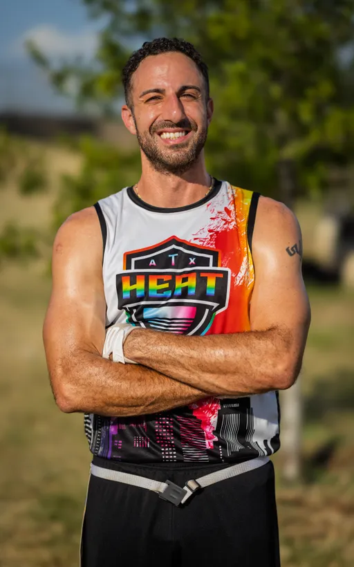 An ATX Heat (Black) player stands on a sunny practice field with arms crossed, wearing a sleeveless ATX Heat jersey with bright orange and pink accents and a flag football belt. Trees blur in the background as the team practices for Gay Bowl 2025.