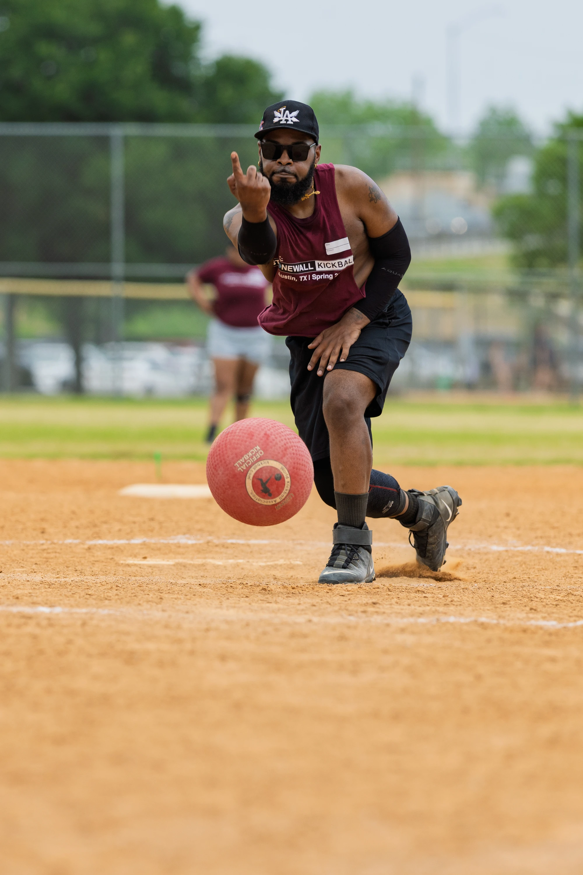 A player wearing a maroon Stonewall Kickball shirt, black shorts, knee brace, sunglasses, and a cap is in the process of pitching a red kickball on a dirt field, with another person visible in the background.