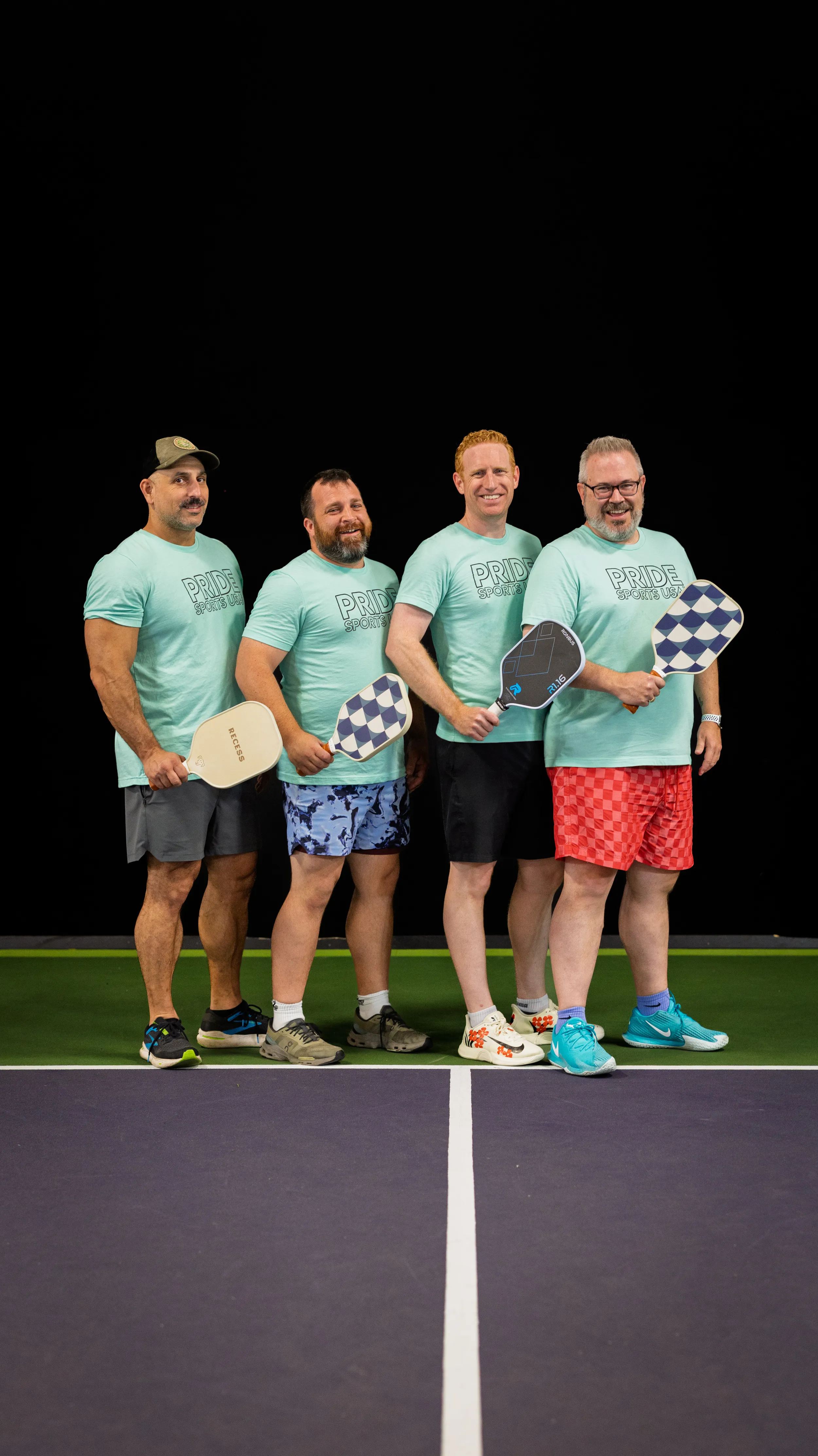 Four players stand side by side on a pickleball court, posing with paddles; they wear matching mint “PRIDE Pickleball” shirts, colorful patterned shorts, and sneakers. A dark backdrop and bright court lines frame the team lineup, with checkered paddles and bold shorts standing out before play.