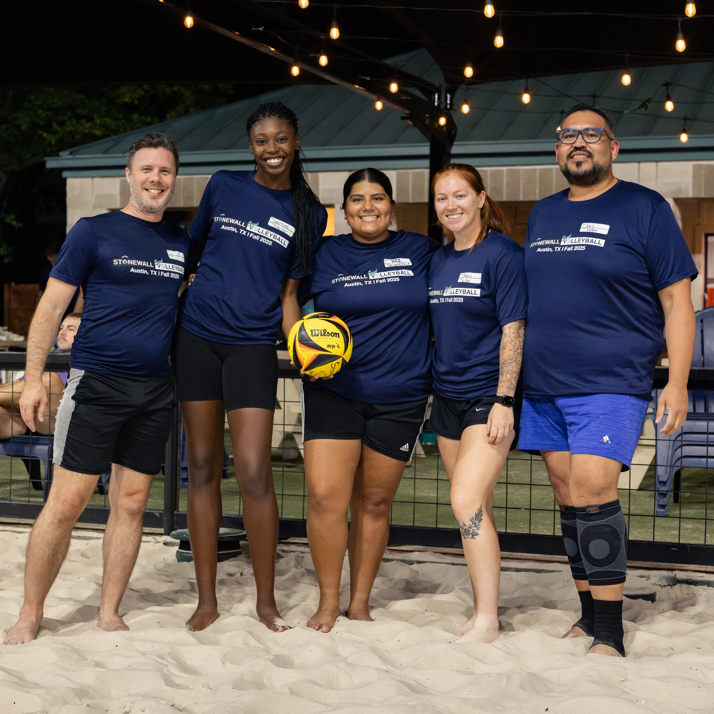 Five players pose together on a sand volleyball court under string lights, wearing matching navy Stonewall Sand Volleyball shirts; the center player holds a yellow Wilson volleyball. A fenced court with a gazebo and benches is in the background, and one player wears a knee brace with everyone in athletic shorts.