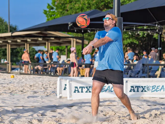 A sand volleyball player in a blue shirt and black shorts prepares to pass a volleyball using a forearm bump, with several people and shaded structures visible in the background on the sand court.